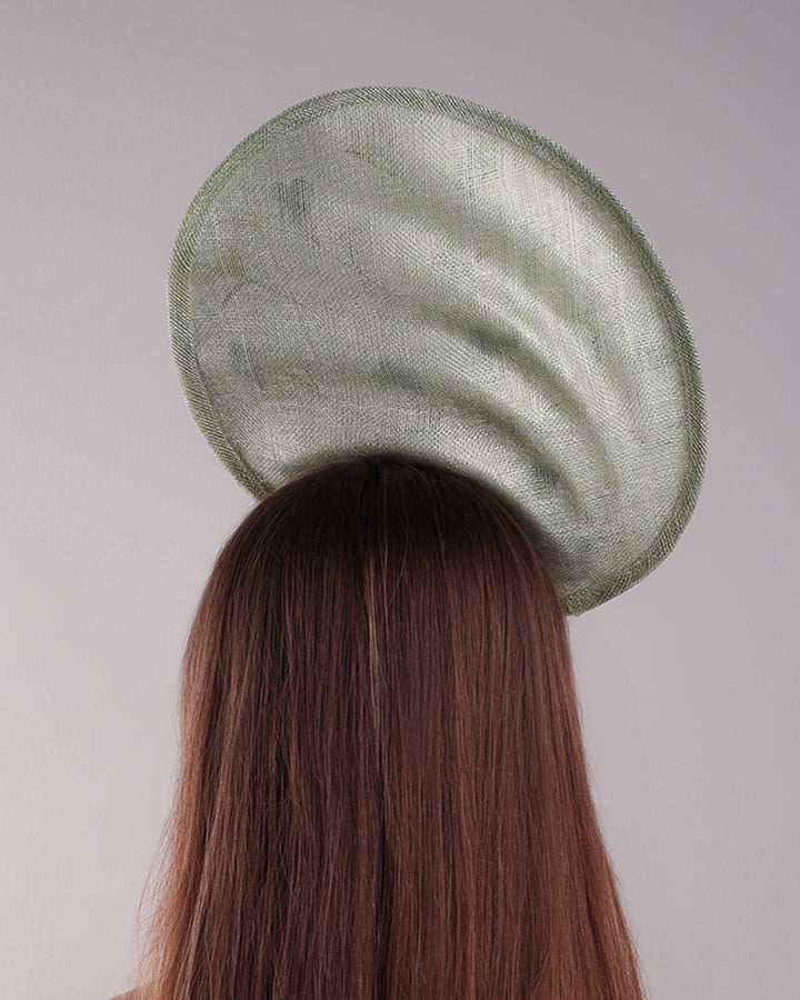 Model is showcasing the back side of light green tall fascinator with curves on the tall side like a sea shell, decorated with multi-colour pressed natural flowers. Fitted to the head with wire headband and elastic.