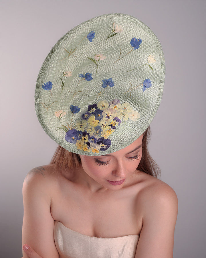 Model is showcasing the top details of the light green tall fascinator with curves on the tall side like a sea shell, decorated with multi-colour pressed natural flowers. Fitted to the head with wire headband and elastic.