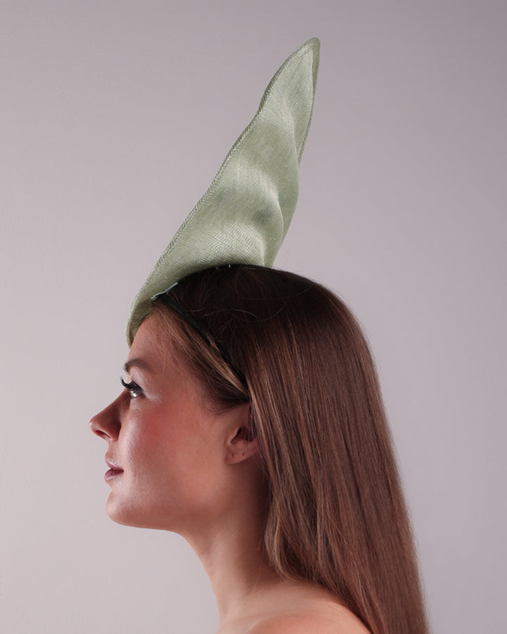 Model is showcasing the left side of light green tall fascinator with curves on the tall side like a sea shell, decorated with multi-colour pressed natural flowers. Fitted to the head with wire headband and elastic.