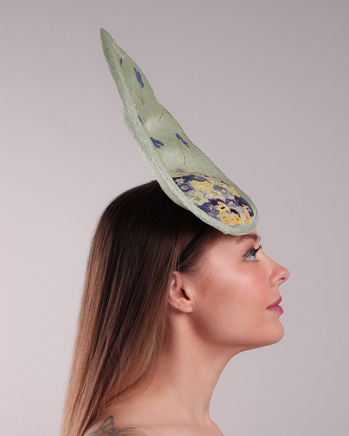 Model is showcasing the right side of light green tall fascinator with curves on the tall side like a sea shell, decorated with multi-colour pressed natural flowers. Fitted to the head with wire headband and elastic.