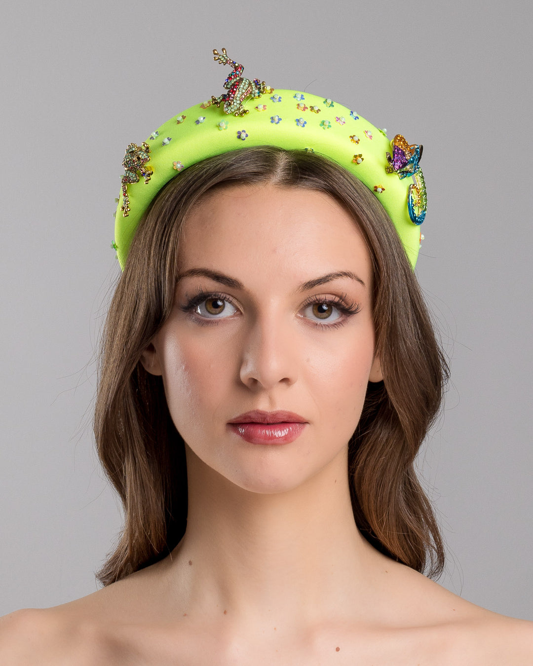 Model is showcasing front side close up of the florescent green high headband embroidered with multi colour flower sequins decorated with two multi colour crystal embellished frog brooches and a multi-colour crystal decorated chameleon. Made by Merve Bayindir Millinery.