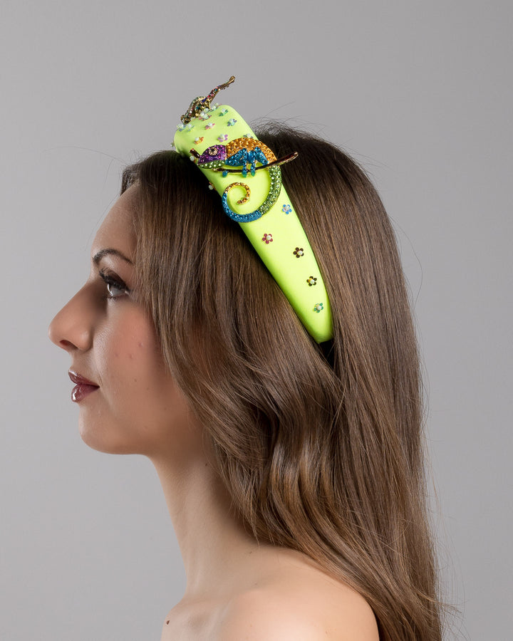 Model is showcasing left side close up of the florescent green high headband embroidered with multi colour flower sequins decorated with two multi colour crystal embellished frog brooches and a multi-colour crystal decorated chameleon. Made by Merve Bayindir Millinery.