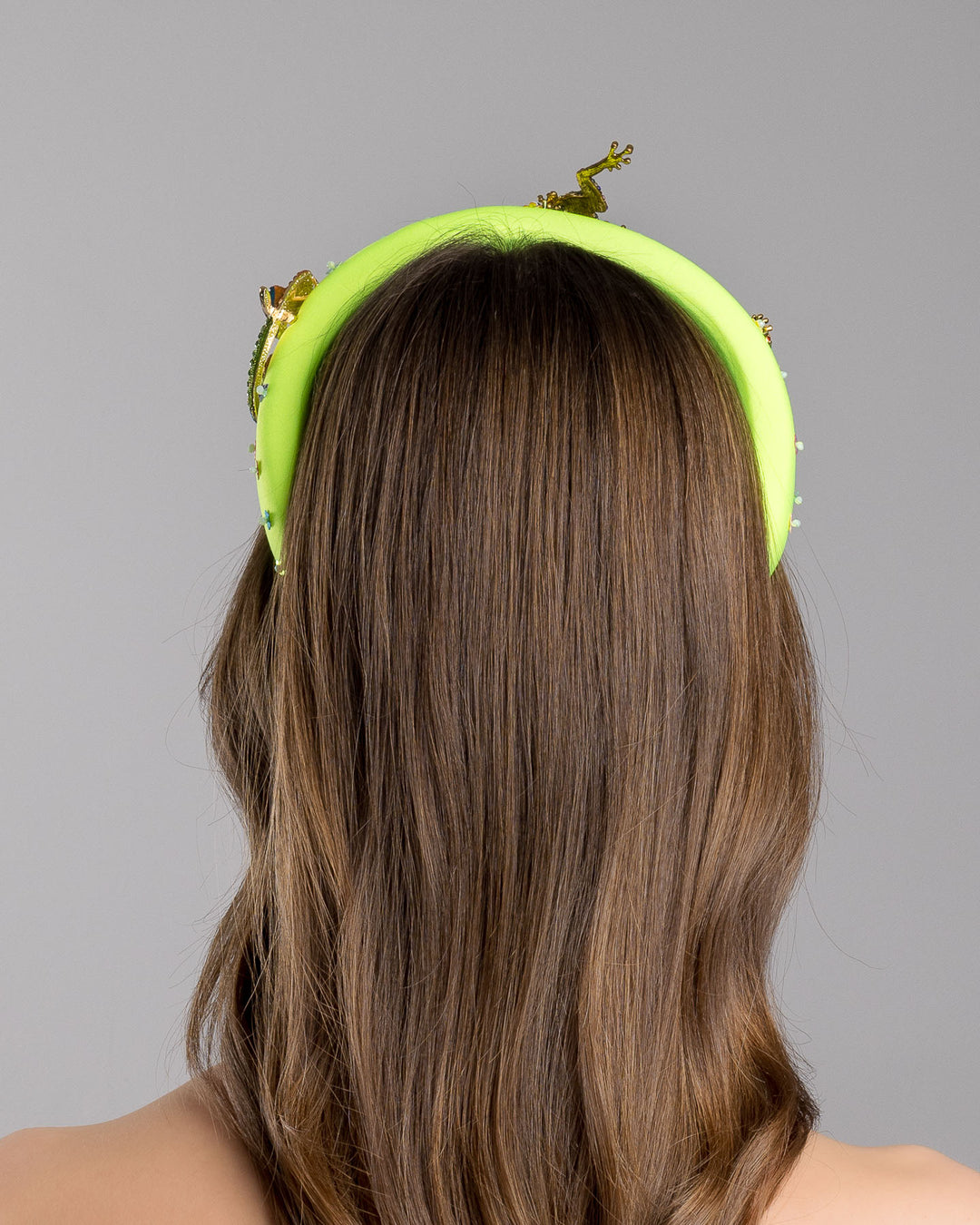 Model is showcasing back side close up of the florescent green high headband embroidered with multi colour flower sequins decorated with two multi colour crystal embellished frog brooches and a multi-colour crystal decorated chameleon. Made by Merve Bayindir Millinery.