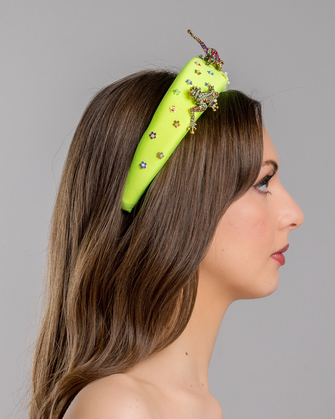 Model is showcasing right side close up of the florescent green high headband embroidered with multi colour flower sequins decorated with two multi colour crystal embellished frog brooches and a multi-colour crystal decorated chameleon. Made by Merve Bayindir Millinery.