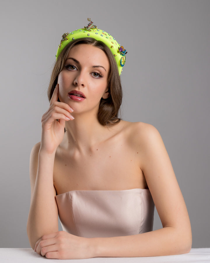 Female model posing wearing Gecko the florescent green high headband embroidered with multi colour flower sequins decorated with two multi colour crystal embellished frog brooches and a multi-colour crystal decorated chameleon. Made by Merve Bayindir Millinery.
