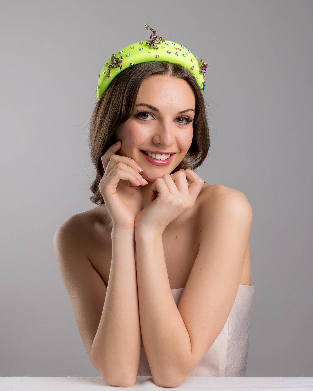 Female model posing wearing Gecko the florescent green high headband embroidered with multi colour flower sequins decorated with two multi colour crystal embellished frog brooches and a multi-colour crystal decorated chameleon. Made by Merve Bayindir Millinery.