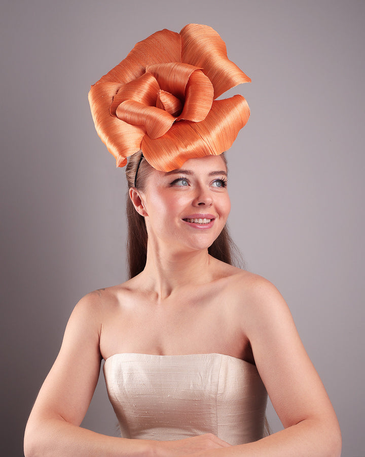 Model is showcasing a front of a large peach orange coloured hand-made silk abaca flower from the lower angle, placed on a sky blue mini saucer. The mini saucer base is fixed on a custom headband and secured with an additional elastic. The peach fascinator is perfect for events like Royal Ascot or Kentucky.