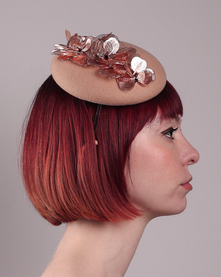 The female model is wearing and showcasing the right side close-up of a camel-coloured button-shaped pillbox fascinator with laser-cut rose-gold coloured metal flowers. Designed by Merve Bayindir.