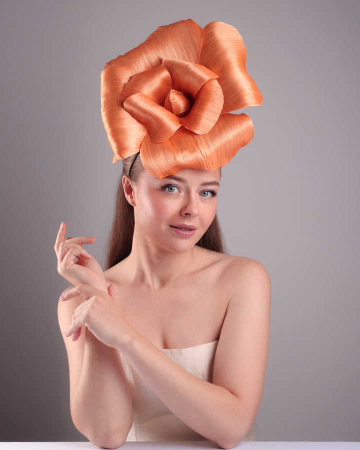 Model is show casing the front of a large peach orange coloured hand-made silk abaca flower, secured with a headband and an additional elastic. The peach fascinator is perfect for events like Royal Ascot or Kentucky.