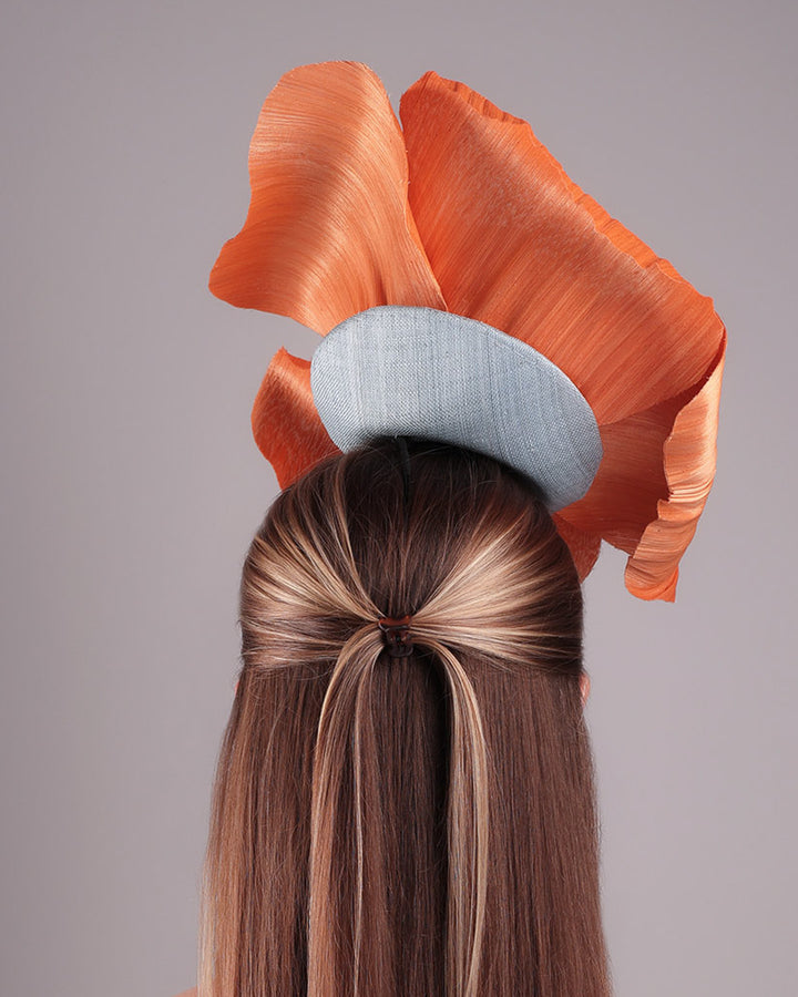 Model is showcasing a back of a large peach orange coloured hand-made silk abaca flower, placed on a sky blue mini saucer. The mini saucer base is fixed on a custom headband and secured with an additional elastic. The peach fascinator is perfect for events like Royal Ascot or Kentucky.