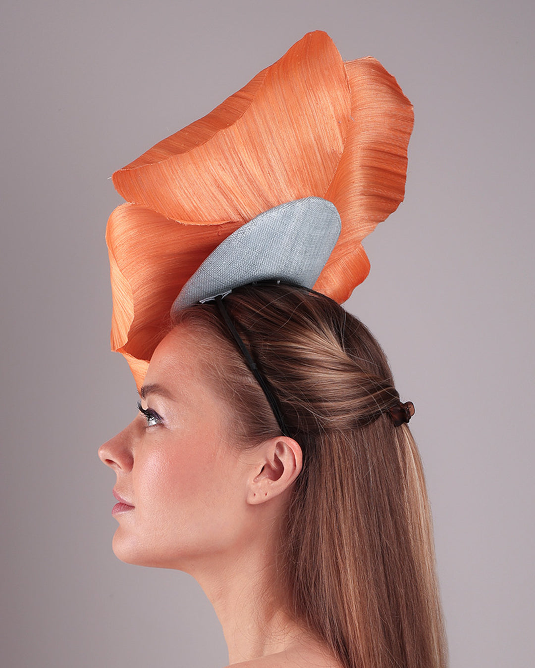 Model is showcasing a left side of a large peach orange coloured hand-made silk abaca flower, placed on a sky blue mini saucer. The mini saucer base is fixed on a custom headband and secured with an additional elastic. The peach fascinator is perfect for events like Royal Ascot or Kentucky.