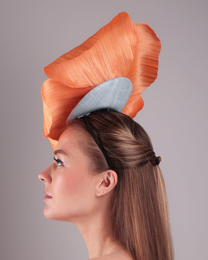 Model is showcasing a left side of a large peach orange coloured hand-made silk abaca flower, placed on a sky blue mini saucer. The mini saucer base is fixed on a custom headband and secured with an additional elastic. The peach fascinator is perfect for events like Royal Ascot or Kentucky.