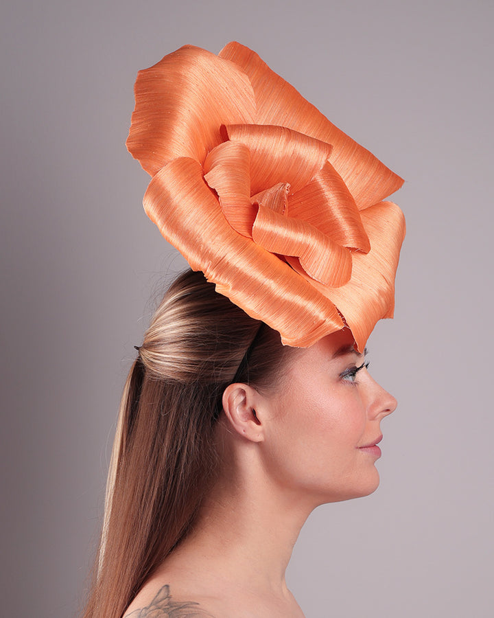 Model is showcasing a right side of a large peach orange coloured hand-made silk abaca flower, placed on a sky blue mini saucer. The mini saucer base is fixed on a custom headband and secured with an additional elastic. The peach fascinator is perfect for events like Royal Ascot or Kentucky.