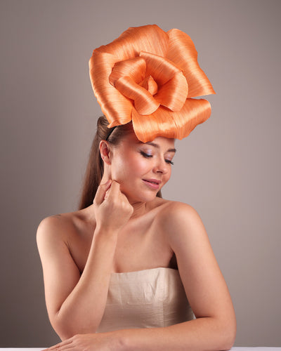 Model is showcasing a front right side of a large peach orange coloured hand-made silk abaca flower, placed on a sky blue mini saucer. The mini saucer base is fixed on a custom headband and secured with an additional elastic. The peach fascinator is perfect for events like Royal Ascot or Kentucky.