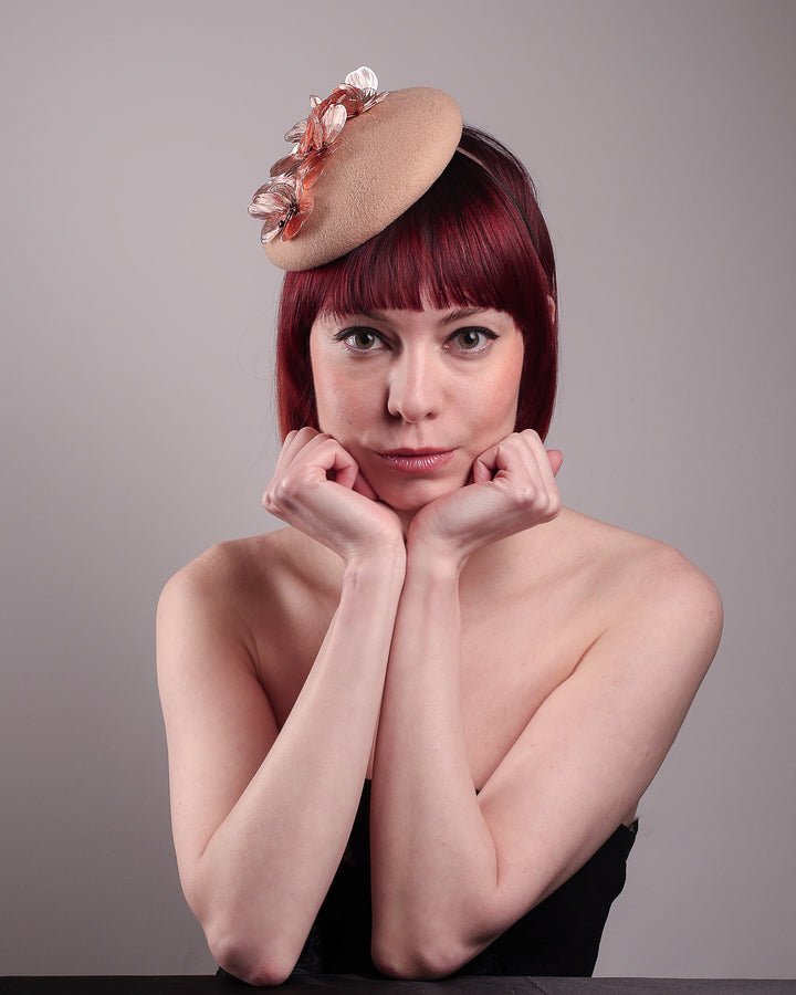 The female model is wearing and showcasing the front style of a camel-coloured button-shaped pillbox fascinator with laser-cut rose-gold coloured metal flowers. Designed by Merve Bayindir.