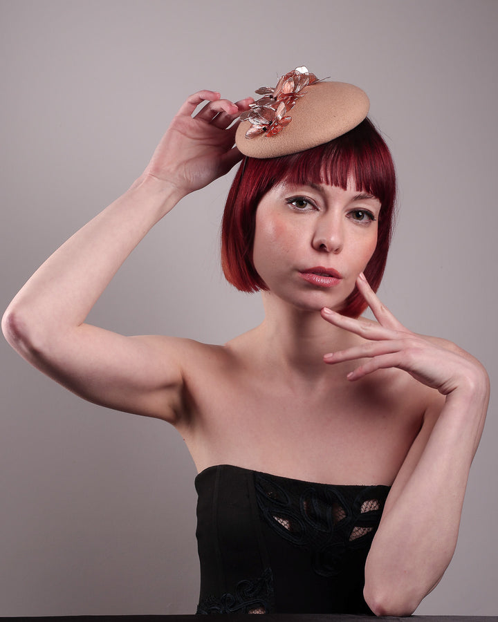 The female model is wearing and showcasing thet front below angle of a camel-coloured button-shaped pillbox fascinator with laser-cut rose-gold coloured metal flowers. Designed by Merve Bayindir.