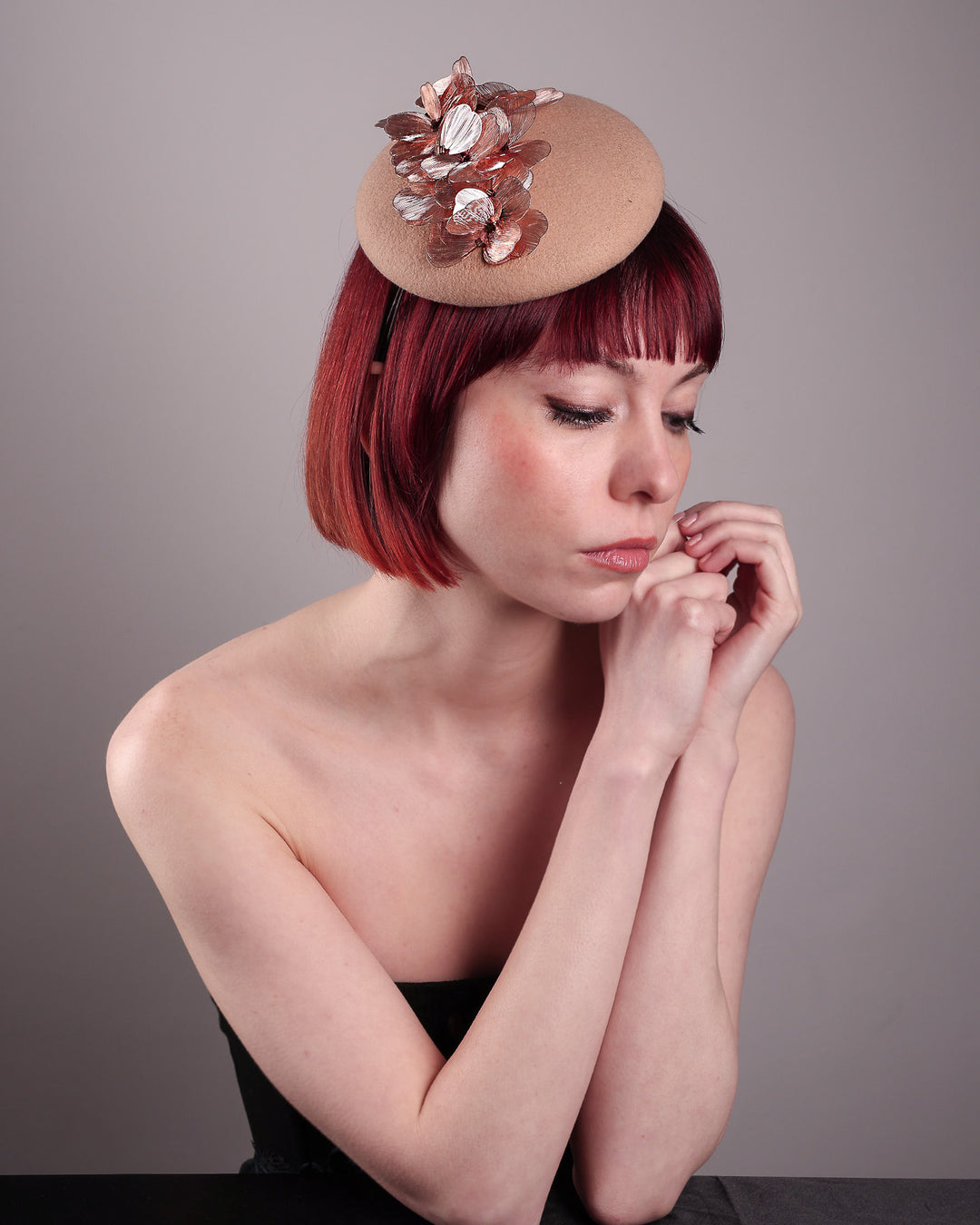 The female model is wearing and showcasing the right front side of a camel-coloured button-shaped pillbox fascinator with laser-cut rose-gold coloured metal flowers. Designed by Merve Bayindir.
