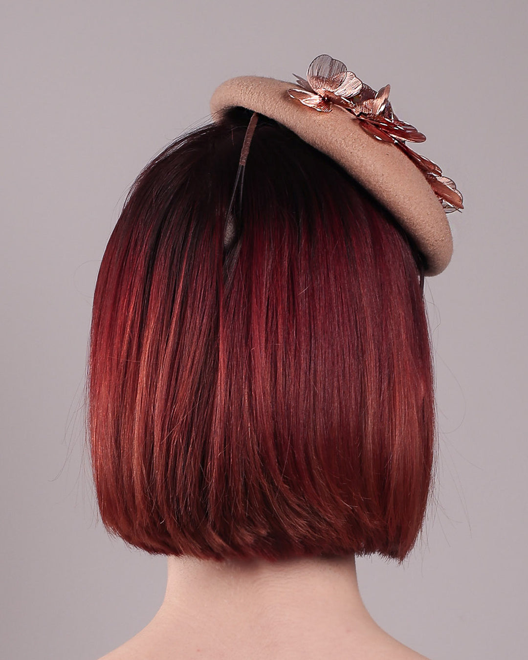The female model is wearing and showcasing the back of a camel-coloured button-shaped pillbox fascinator with laser-cut rose-gold coloured metal flowers. Designed by Merve Bayindir.