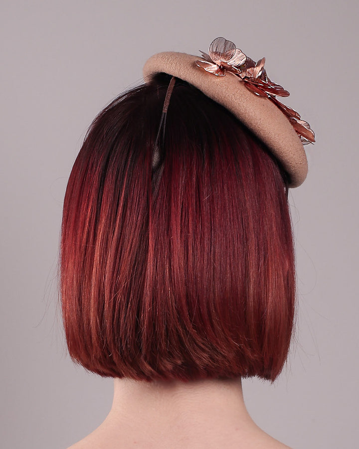 The female model is wearing and showcasing the back of a camel-coloured button-shaped pillbox fascinator with laser-cut rose-gold coloured metal flowers. Designed by Merve Bayindir.