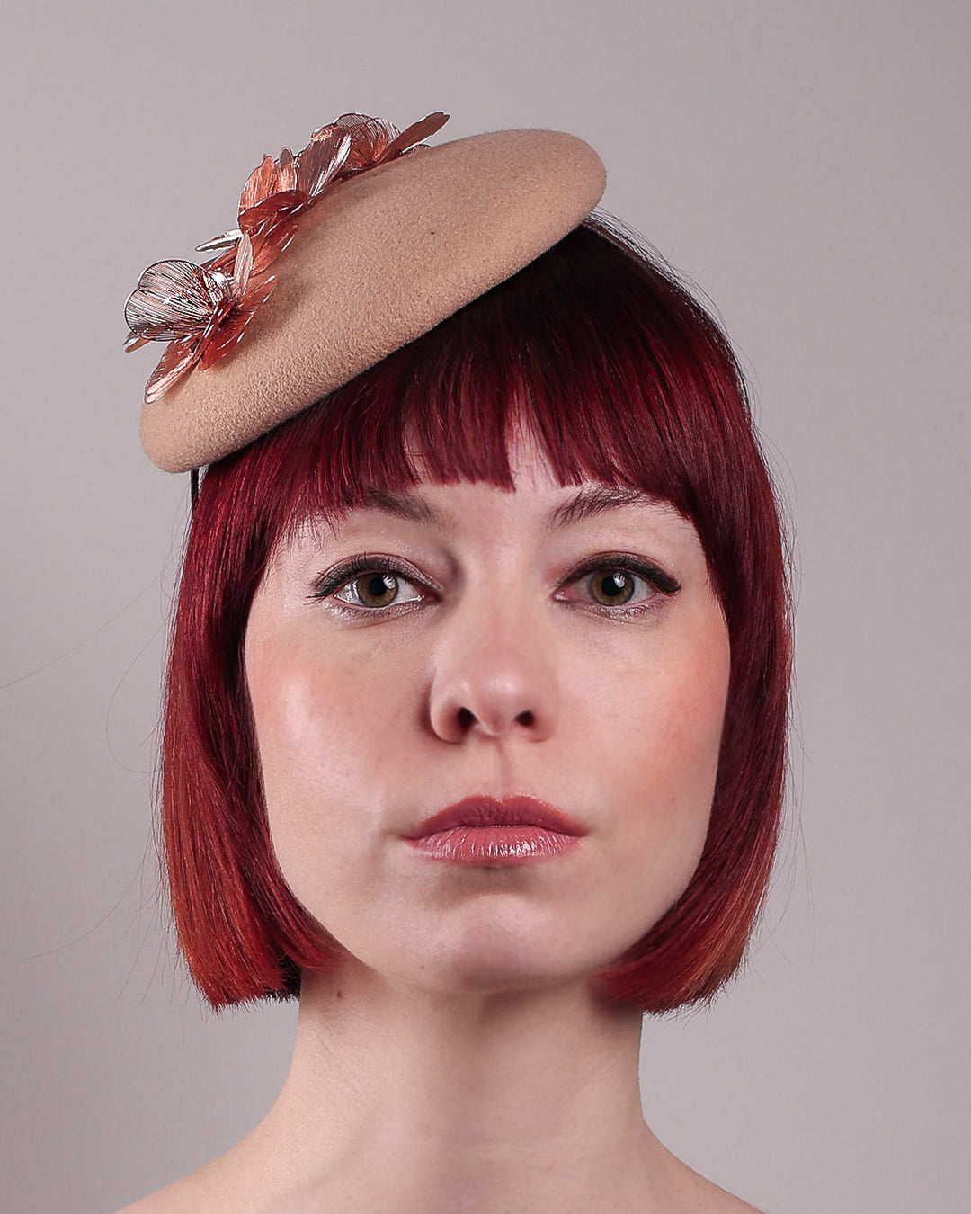 The female model is wearing and showcasing the front close-up of a camel-coloured button-shaped pillbox fascinator with laser-cut rose-gold coloured metal flowers. Designed by Merve Bayindir.
