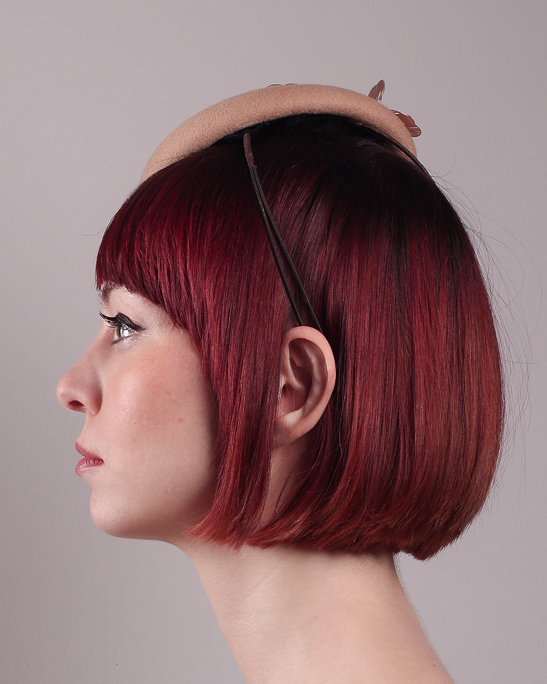 The female model is wearing and showcasing the left side close-up of a camel-coloured button-shaped pillbox fascinator with laser-cut rose-gold coloured metal flowers. Designed by Merve Bayindir