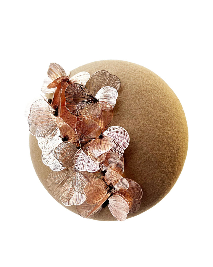 Still life picture of a camel-coloured button-shaped pillbox fascinator with laser-cut rose-gold coloured metal flowers. Designed by Merve Bayindir.