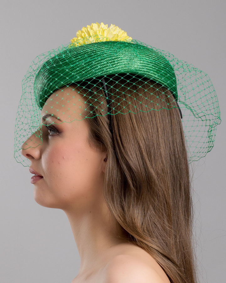 Model is showcasing the left side close up of green summer beret tilted to the right side of the head, the vibrant headpiece is rounded with a delicate emerald green veil and completed with a bright yellow pompom flower on top. Designed and made by Merve Bayindir millinery.