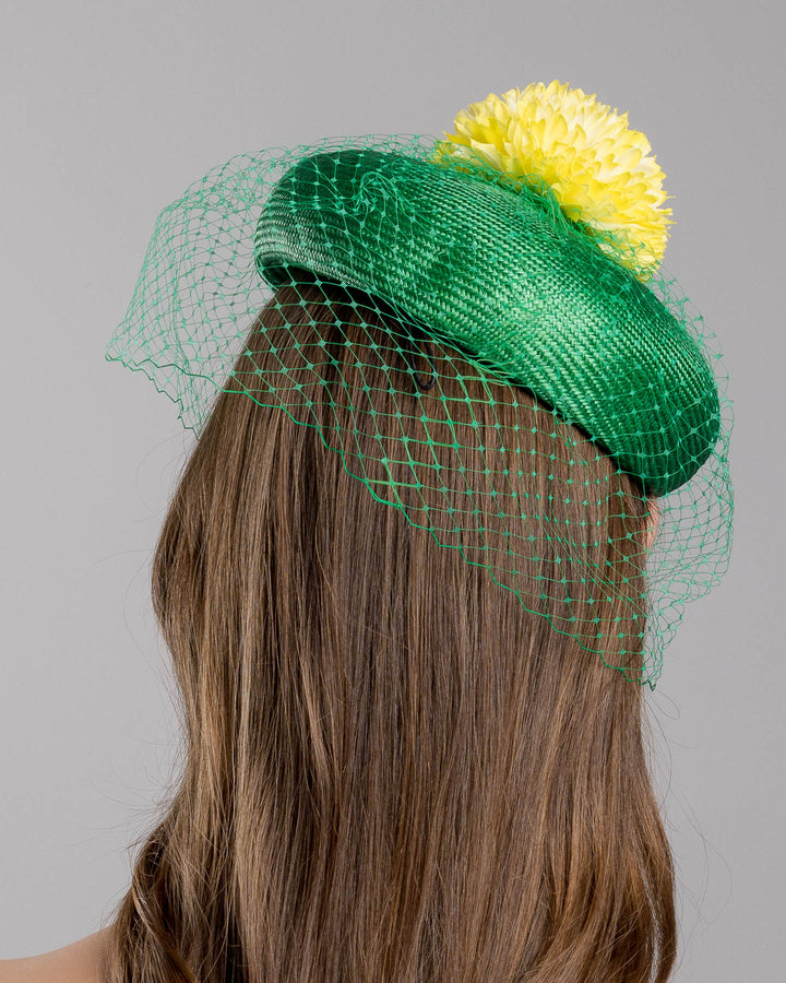 Model is showcasing the back side close up of green summer beret tilted to the right side of the head, the vibrant headpiece is rounded with a delicate emerald green veil and completed with a bright yellow pompom flower on top. Designed and made by Merve Bayindir millinery.