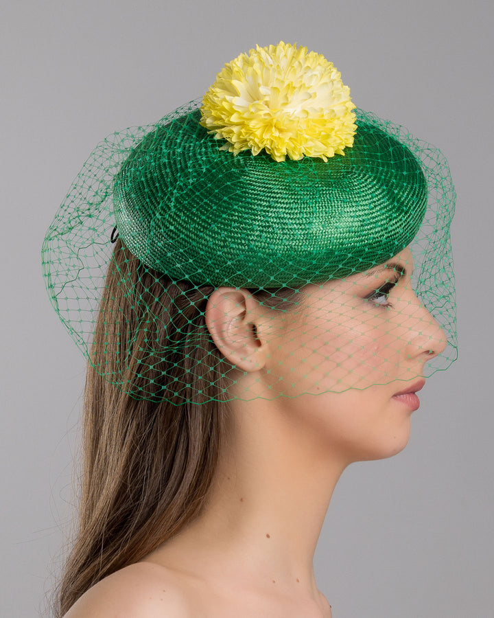 Model is showcasing the right side close up of green summer beret tilted to the right side of the head, the vibrant headpiece is rounded with a delicate emerald green veil and completed with a bright yellow pompom flower on top. Designed and made by Merve Bayindir millinery.