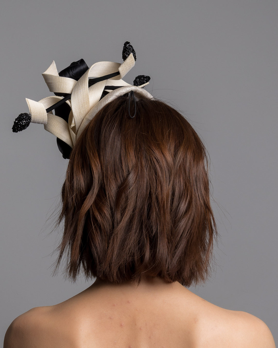 The model is showcasing the back side close up of the grade sinamay button shaped white fascinator decorated with white Jinsin Swirls and custom made black American Sateen Cala flower. Designed and made by Merve Bayindir Millinery.

