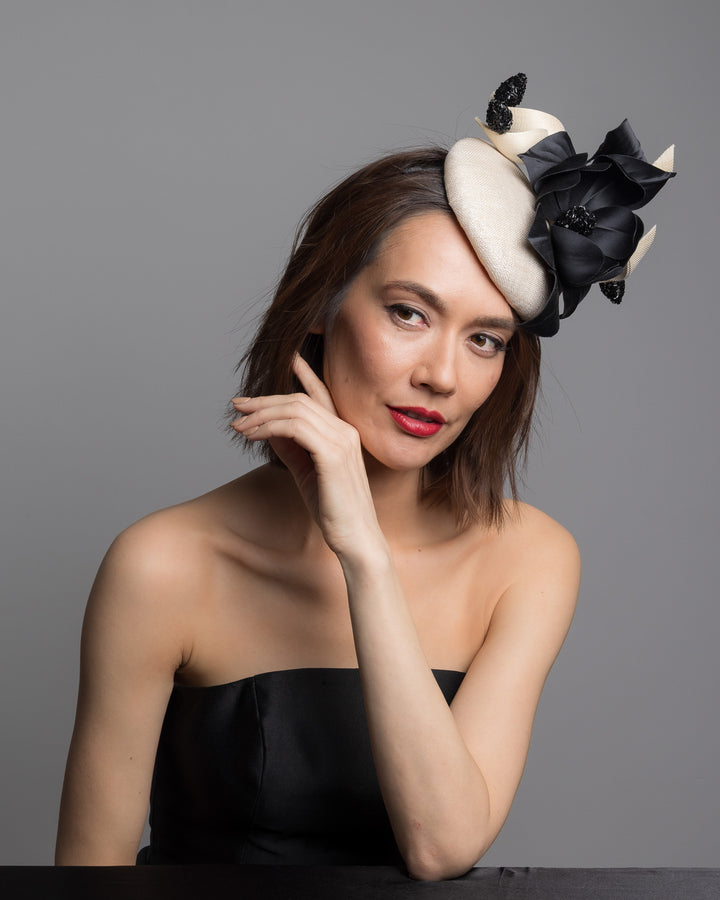 The female model is posing, wearing 
Guinea,the grade sinamay button shaped white fascinator decorated with white Jinsin Swirls and custom made black American Sateen Cala flower. Designed and made by Merve Bayindir Millinery.

