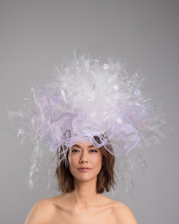 The model is showcasing the front side close up of the avant-garde statement headpiece composed of an abundant explosion of airy white and soft lilac fibers. The design features layered, cloud-like textures with wispy strands, fine feather-like elements, and sheer tulle interwoven throughout. Its voluminous, sculptural silhouette rises dramatically upward and outward, creating a light, ethereal halo effect around the head with a soft pastel sheen. Designed and made by Merve Bayindir Millinery.
