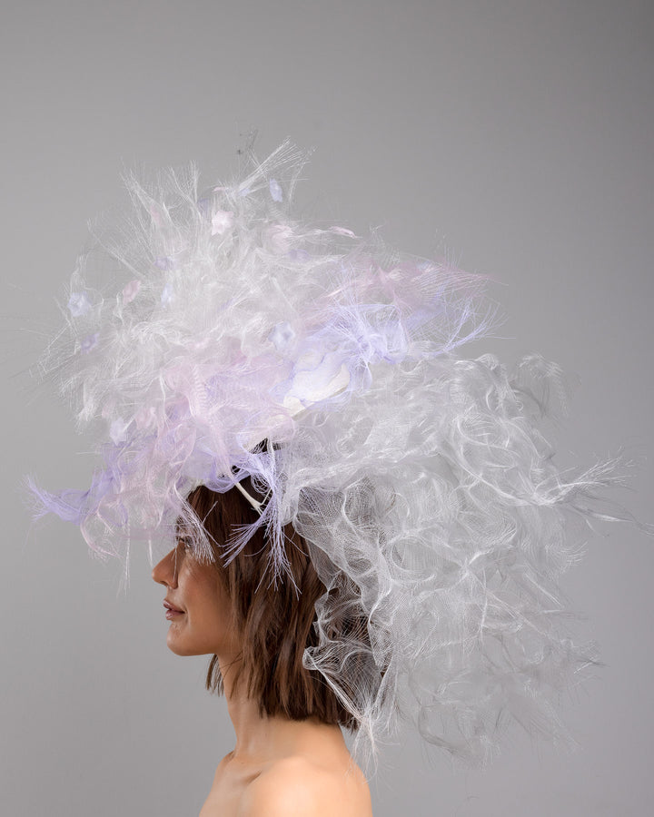 The model is showcasing the left side close up of the avant-garde statement headpiece composed of an abundant explosion of airy white and soft lilac fibers. The design features layered, cloud-like textures with wispy strands, fine feather-like elements, and sheer tulle interwoven throughout. Its voluminous, sculptural silhouette rises dramatically upward and outward, creating a light, ethereal halo effect around the head with a soft pastel sheen. Designed and made by Merve Bayindir Millinery.
