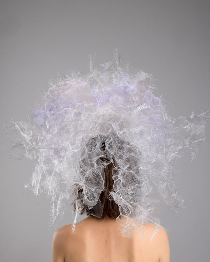 The model is showcasing the back side close up of the avant-garde statement headpiece composed of an abundant explosion of airy white and soft lilac fibers. The design features layered, cloud-like textures with wispy strands, fine feather-like elements, and sheer tulle interwoven throughout. Its voluminous, sculptural silhouette rises dramatically upward and outward, creating a light, ethereal halo effect around the head with a soft pastel sheen. Designed and made by Merve Bayindir Millinery.
