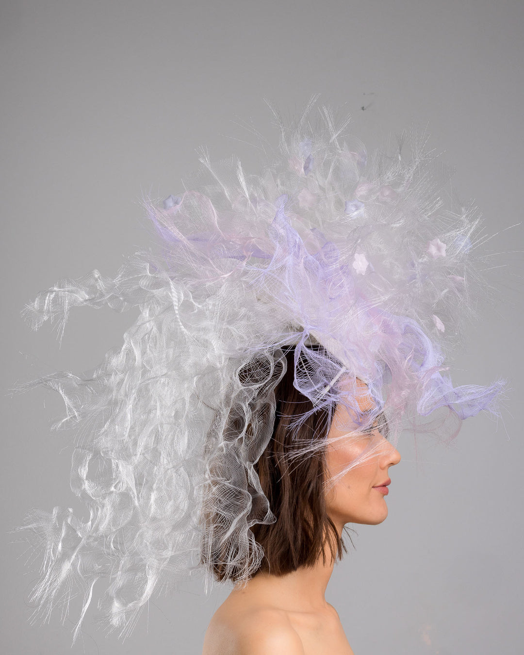 The model is showcasing the right side close up of the avant-garde statement headpiece composed of an abundant explosion of airy white and soft lilac fibers. The design features layered, cloud-like textures with wispy strands, fine feather-like elements, and sheer tulle interwoven throughout. Its voluminous, sculptural silhouette rises dramatically upward and outward, creating a light, ethereal halo effect around the head with a soft pastel sheen. Designed and made by Merve Bayindir Millinery.
