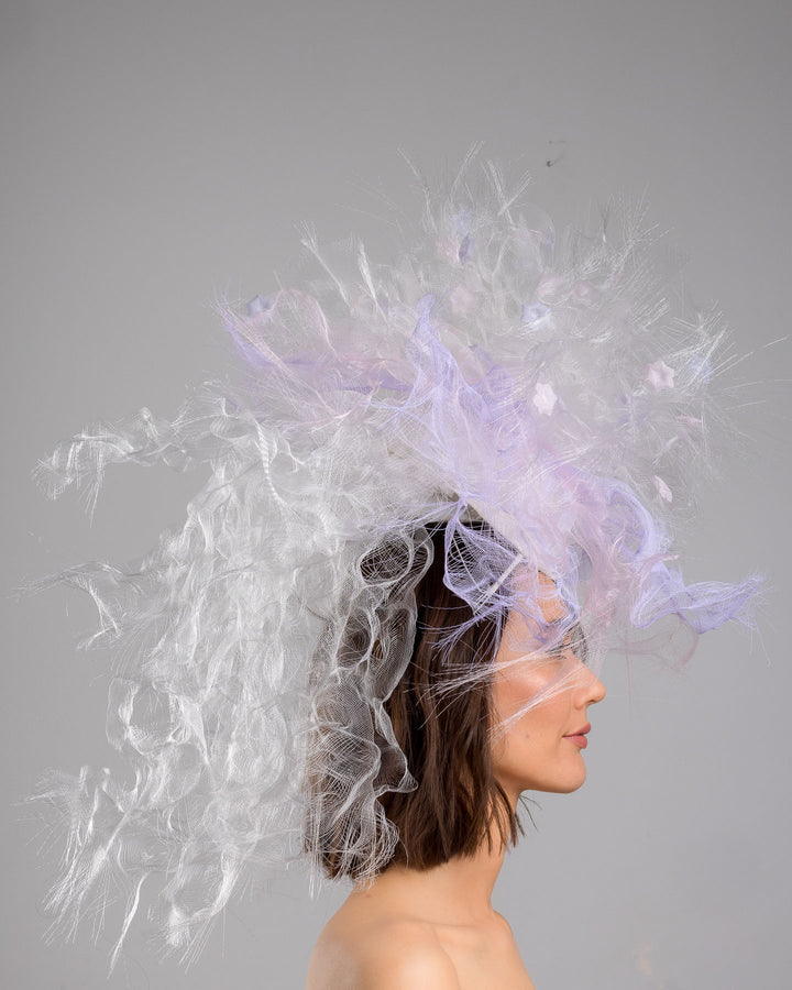 The model is showcasing the right side close up of the avant-garde statement headpiece composed of an abundant explosion of airy white and soft lilac fibers. The design features layered, cloud-like textures with wispy strands, fine feather-like elements, and sheer tulle interwoven throughout. Its voluminous, sculptural silhouette rises dramatically upward and outward, creating a light, ethereal halo effect around the head with a soft pastel sheen. Designed and made by Merve Bayindir Millinery.
