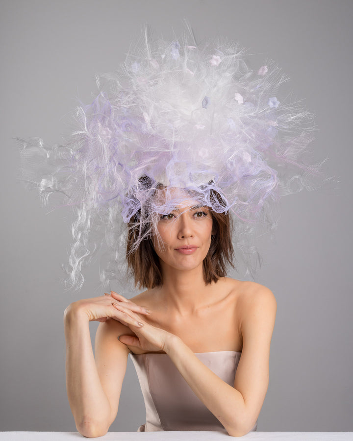 The female model is posing, wearing
Haloir, the avant-garde statement headpiece composed of an abundant explosion of airy white and soft lilac fibers. The design features layered, cloud-like textures with wispy strands, fine feather-like elements, and sheer tulle interwoven throughout. Its voluminous, sculptural silhouette rises dramatically upward and outward, creating a light, ethereal halo effect around the head with a soft pastel sheen. Designed and made by Merve Bayindir Millinery.