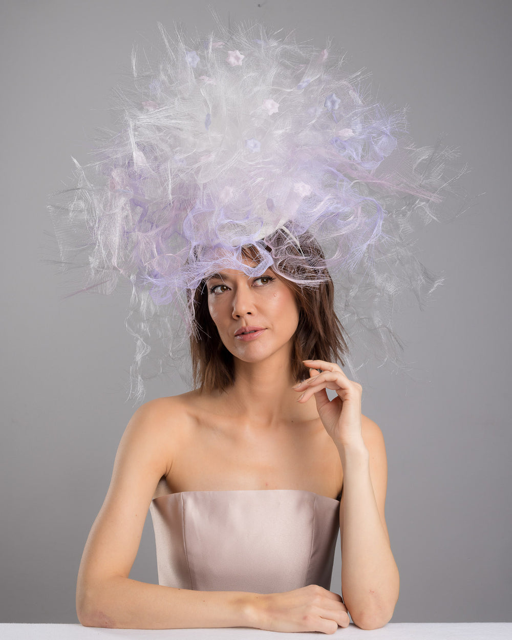 The female model is posing, wearing
Haloir, the avant-garde statement headpiece composed of an abundant explosion of airy white and soft lilac fibers. The design features layered, cloud-like textures with wispy strands, fine feather-like elements, and sheer tulle interwoven throughout. Its voluminous, sculptural silhouette rises dramatically upward and outward, creating a light, ethereal halo effect around the head with a soft pastel sheen. Designed and made by Merve Bayindir Millinery.