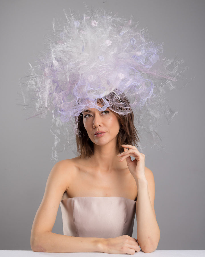 The female model is posing, wearing
Haloir, the avant-garde statement headpiece composed of an abundant explosion of airy white and soft lilac fibers. The design features layered, cloud-like textures with wispy strands, fine feather-like elements, and sheer tulle interwoven throughout. Its voluminous, sculptural silhouette rises dramatically upward and outward, creating a light, ethereal halo effect around the head with a soft pastel sheen. Designed and made by Merve Bayindir Millinery.