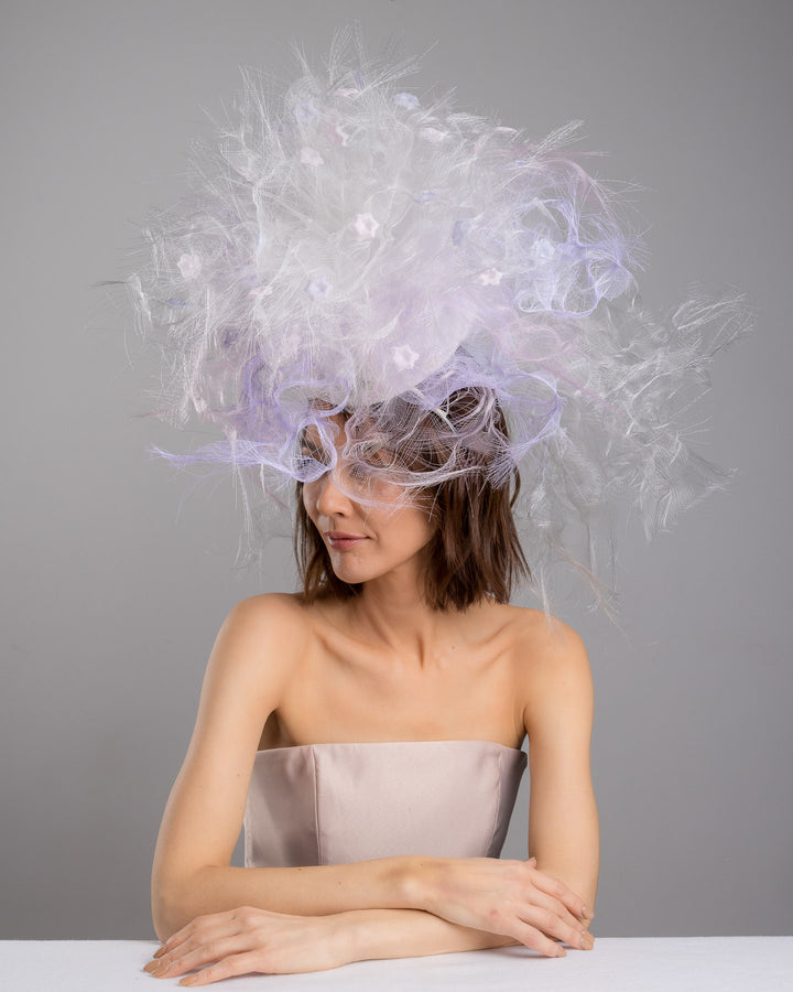 The female model is posing, wearing
Haloir, the avant-garde statement headpiece composed of an abundant explosion of airy white and soft lilac fibers. The design features layered, cloud-like textures with wispy strands, fine feather-like elements, and sheer tulle interwoven throughout. Its voluminous, sculptural silhouette rises dramatically upward and outward, creating a light, ethereal halo effect around the head with a soft pastel sheen. Designed and made by Merve Bayindir Millinery.