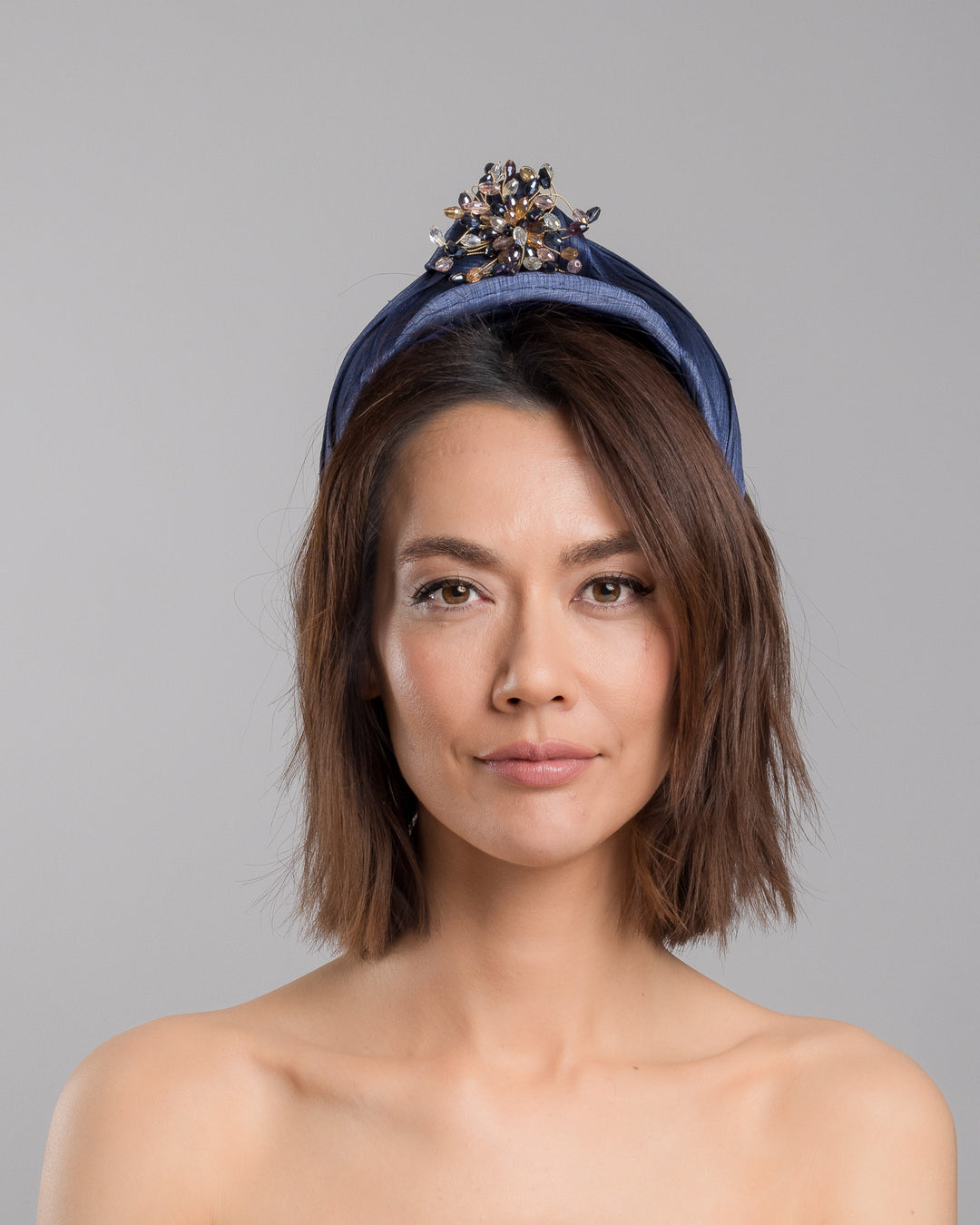 The model is showcasing the front side close up of the navy blue structured half-hat in textured fabric, featuring a sculptural twisted silhouette and embellished with a statement crystal burst brooch in gold, smoky, and blush tones. Designed and made by Merve Bayindir Millinery.
