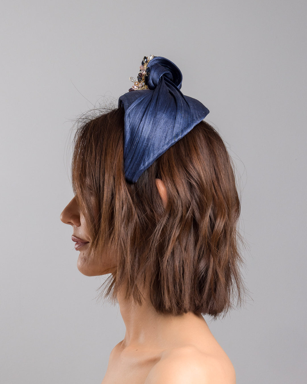The model is showcasing the left side close up of the navy blue structured half-hat in textured fabric, featuring a sculptural twisted silhouette and embellished with a statement crystal burst brooch in gold, smoky, and blush tones. Designed and made by Merve Bayindir Millinery.
