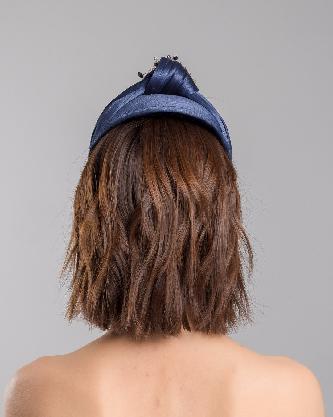 The model is showcasing the back side close up of the navy blue structured half-hat in textured fabric, featuring a sculptural twisted silhouette and embellished with a statement crystal burst brooch in gold, smoky, and blush tones. Designed and made by Merve Bayindir Millinery.
