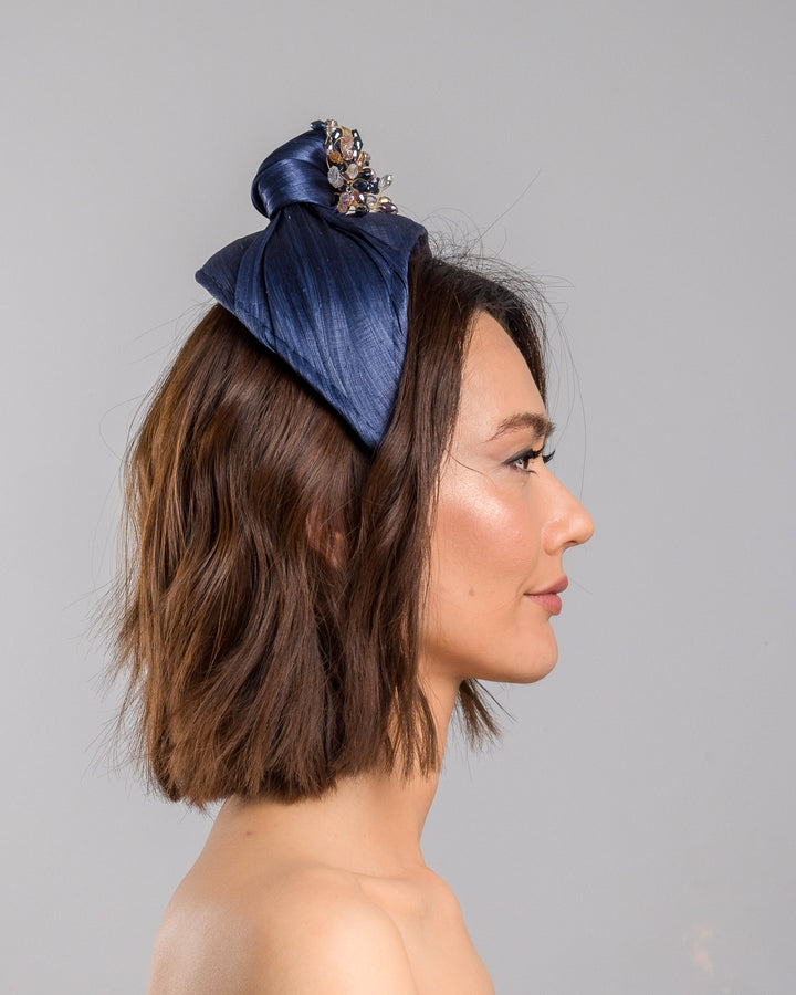 The model is showcasing the right side close up of the navy blue structured half-hat in textured fabric, featuring a sculptural twisted silhouette and embellished with a statement crystal burst brooch in gold, smoky, and blush tones. Designed and made by Merve Bayindir Millinery.
