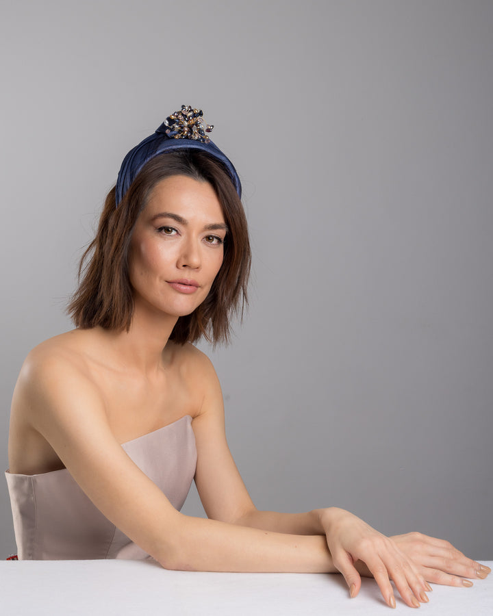 The female model is posing, wearing
Honor, the navy blue structured half-hat in textured fabric, featuring a sculptural twisted silhouette and embellished with a statement crystal burst brooch in gold, smoky, and blush tones. Designed and made by Merve Bayindir Millinery.
