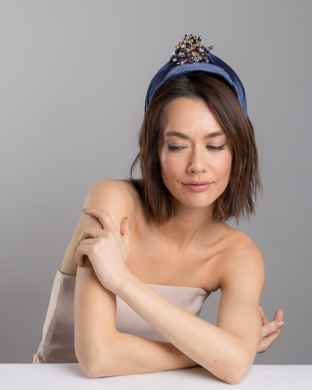 The female model is posing, wearing
Honor, the navy blue structured half-hat in textured fabric, featuring a sculptural twisted silhouette and embellished with a statement crystal burst brooch in gold, smoky, and blush tones. Designed and made by Merve Bayindir Millinery.
