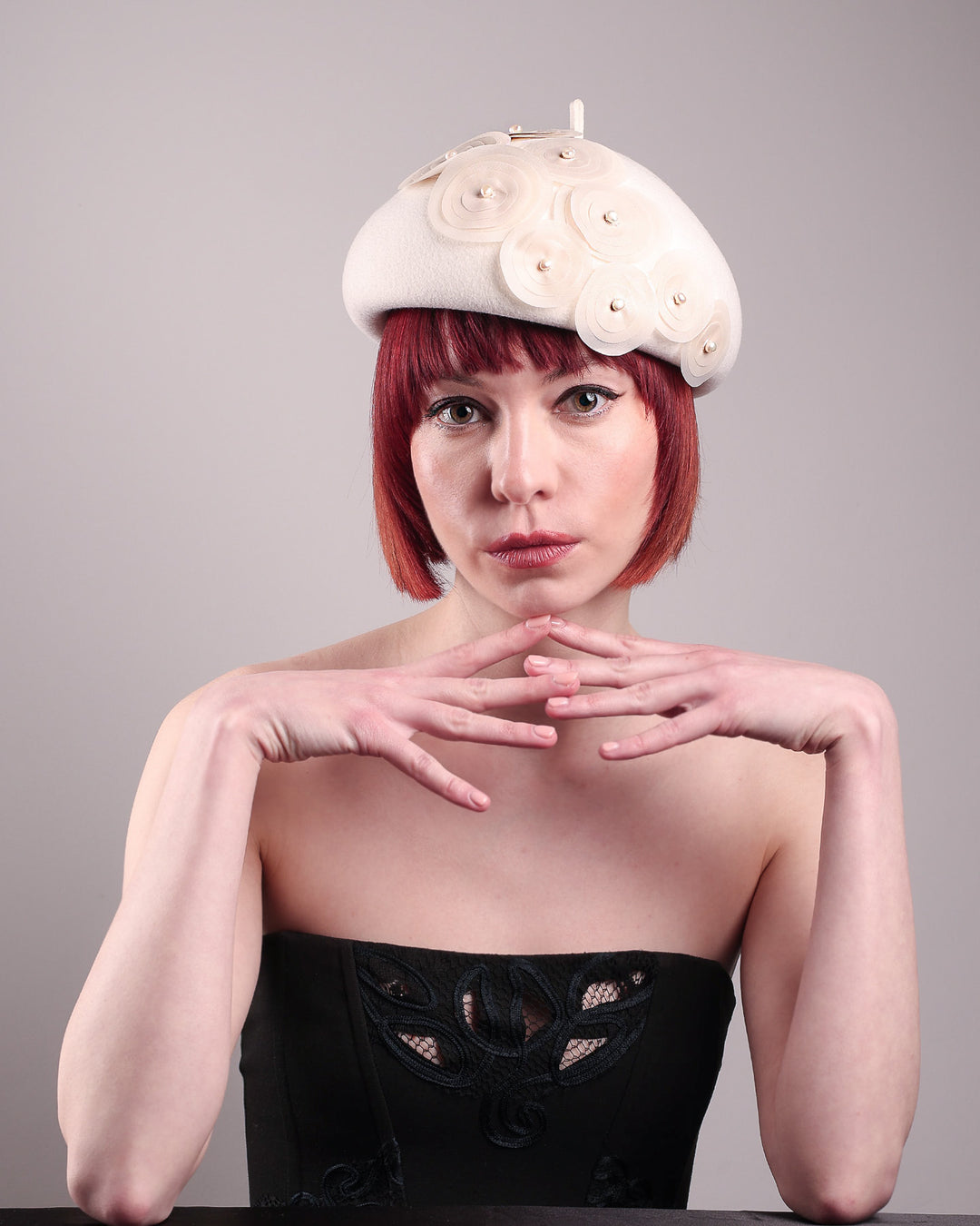 The model is showcasing the fornt view of the Heaven pillbox beret hat, crafted in soft white felt and adorned with swirling ivory appliqué rosettes of different sizes and layers, each hand-finished with a delicate pearl. Designed by Merve Bayindir.