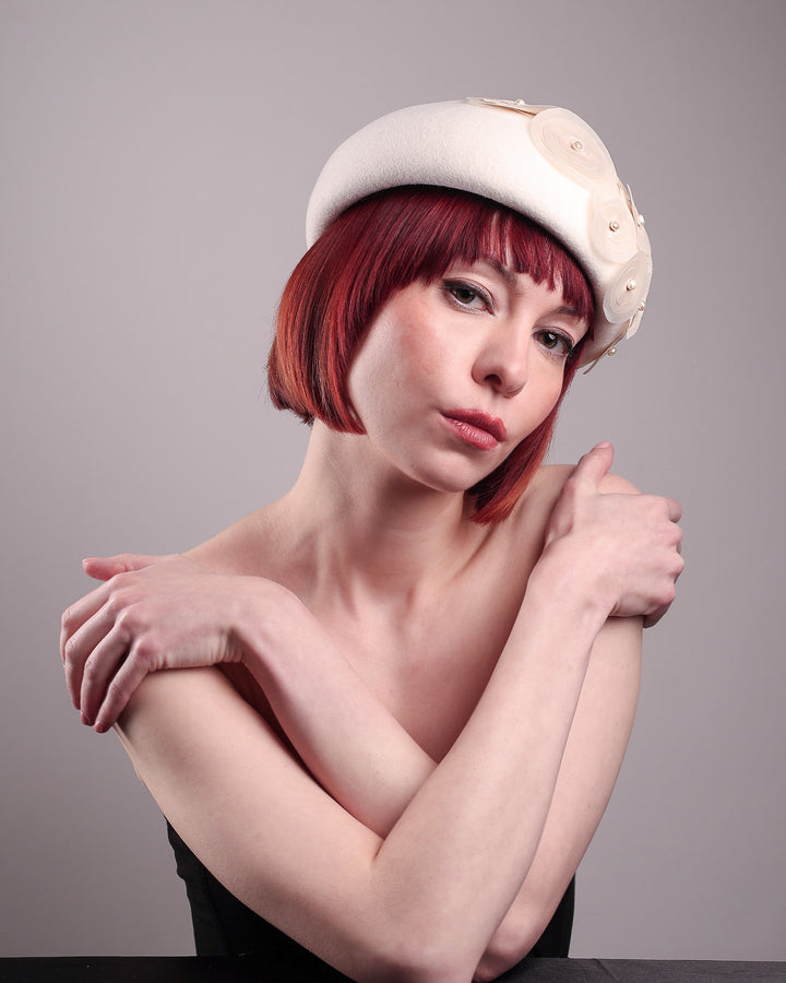 The model is showcasing below right side angle of the Heaven pillbox beret hat, crafted in soft white felt and adorned with swirling ivory appliqué rosettes of different sizes and layers, each hand-finished with a delicate pearl. Designed by Merve Bayindir.