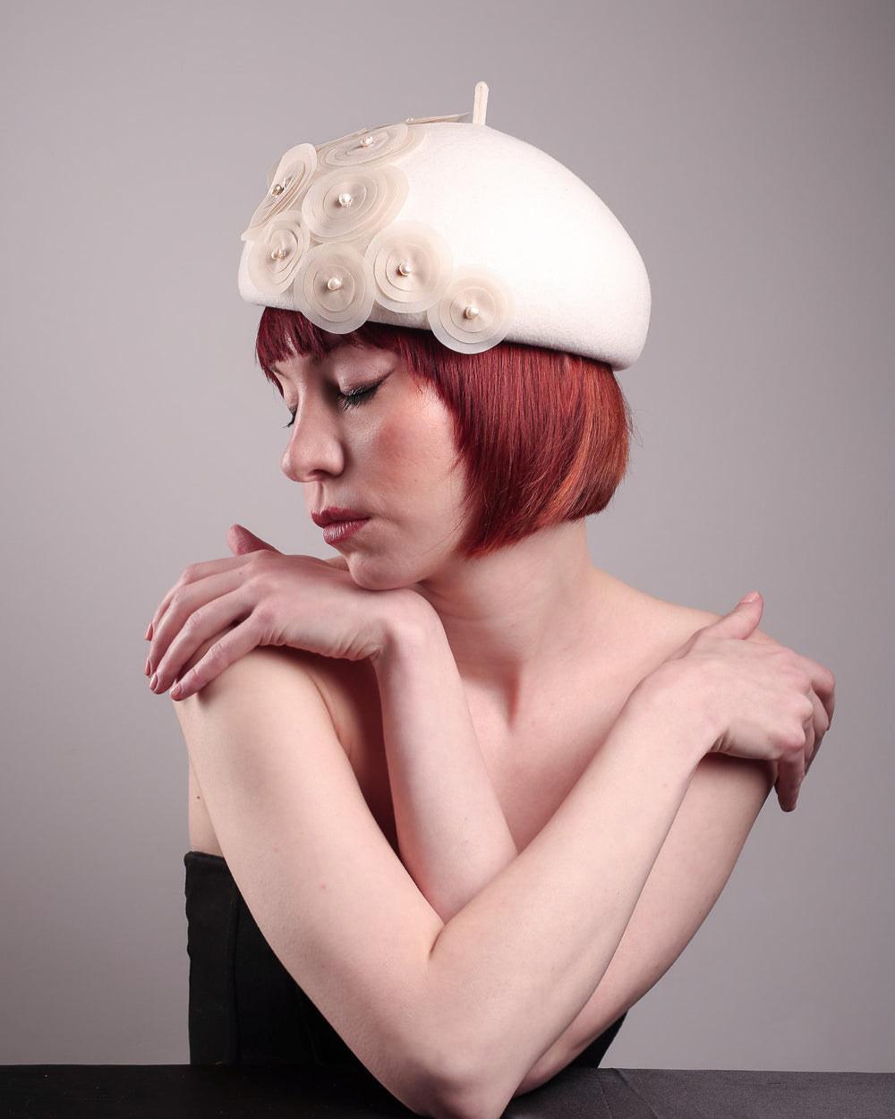 The model is showcasing the left side of the Heaven pillbox beret hat, crafted in soft white felt and adorned with swirling ivory appliqué rosettes of different sizes and layers, each hand-finished with a delicate pearl. Designed by Merve Bayindir.
