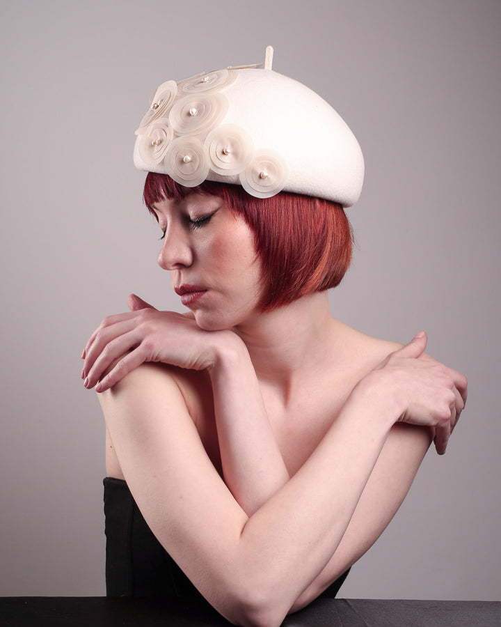 The model is showcasing the left side of the Heaven pillbox beret hat, crafted in soft white felt and adorned with swirling ivory appliqué rosettes of different sizes and layers, each hand-finished with a delicate pearl. Designed by Merve Bayindir.